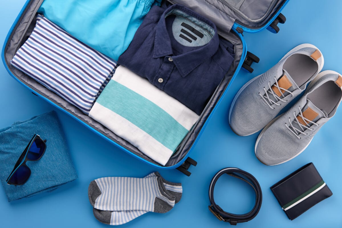 Packing men's summer clothes and accessories in blue travel suitcase on blue background, top view