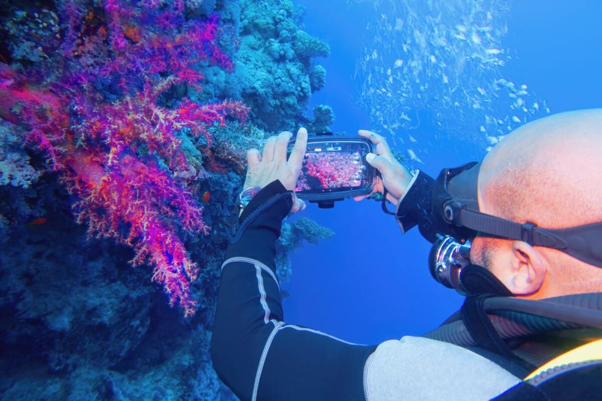 Man scuba diver using smartphone underwater housing when shooting coral reef scenery. Modern smartphone technology.