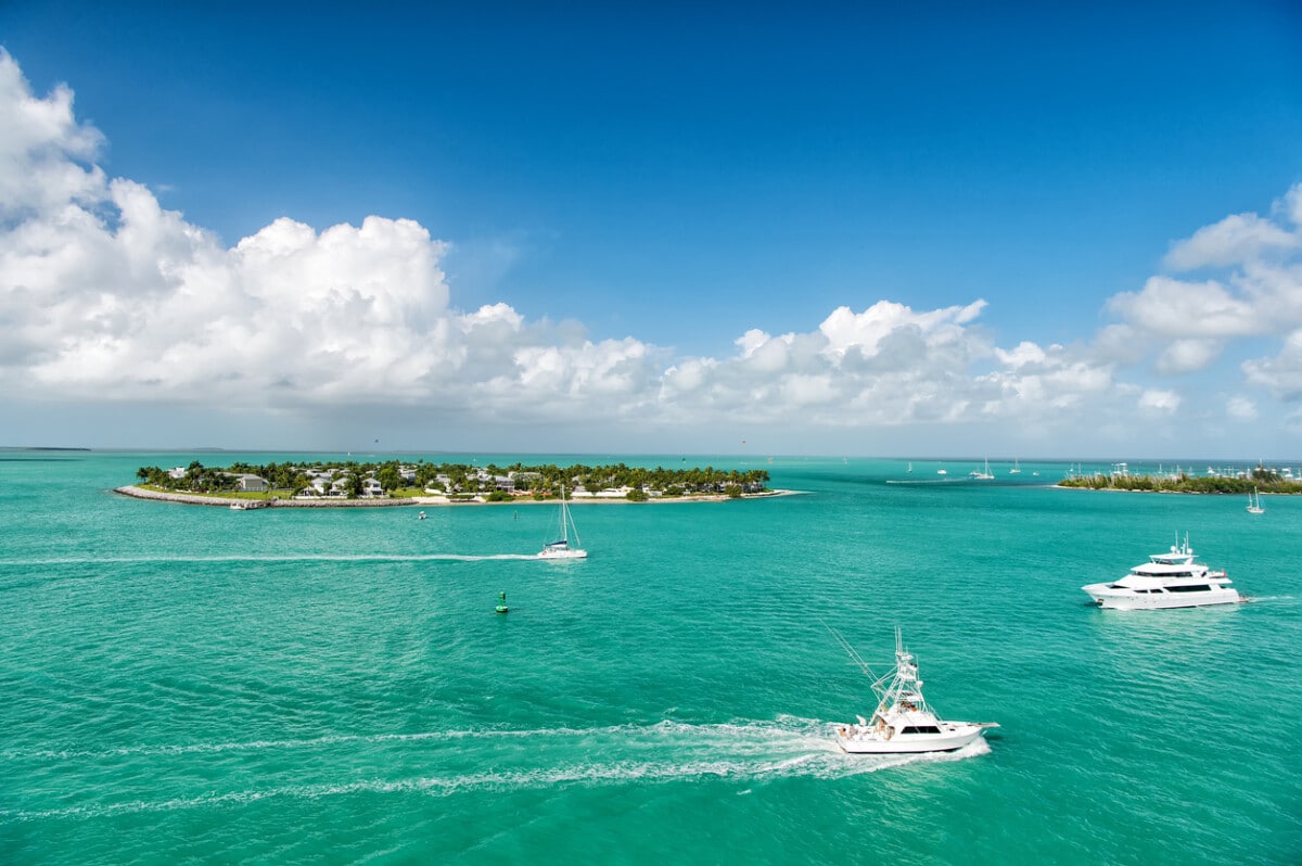 cruise touristic boats or yachts floating by island with houses and green trees on turquoise water and blue cloudy sky, yachting and isle life around beautiful Key West Florida, USA